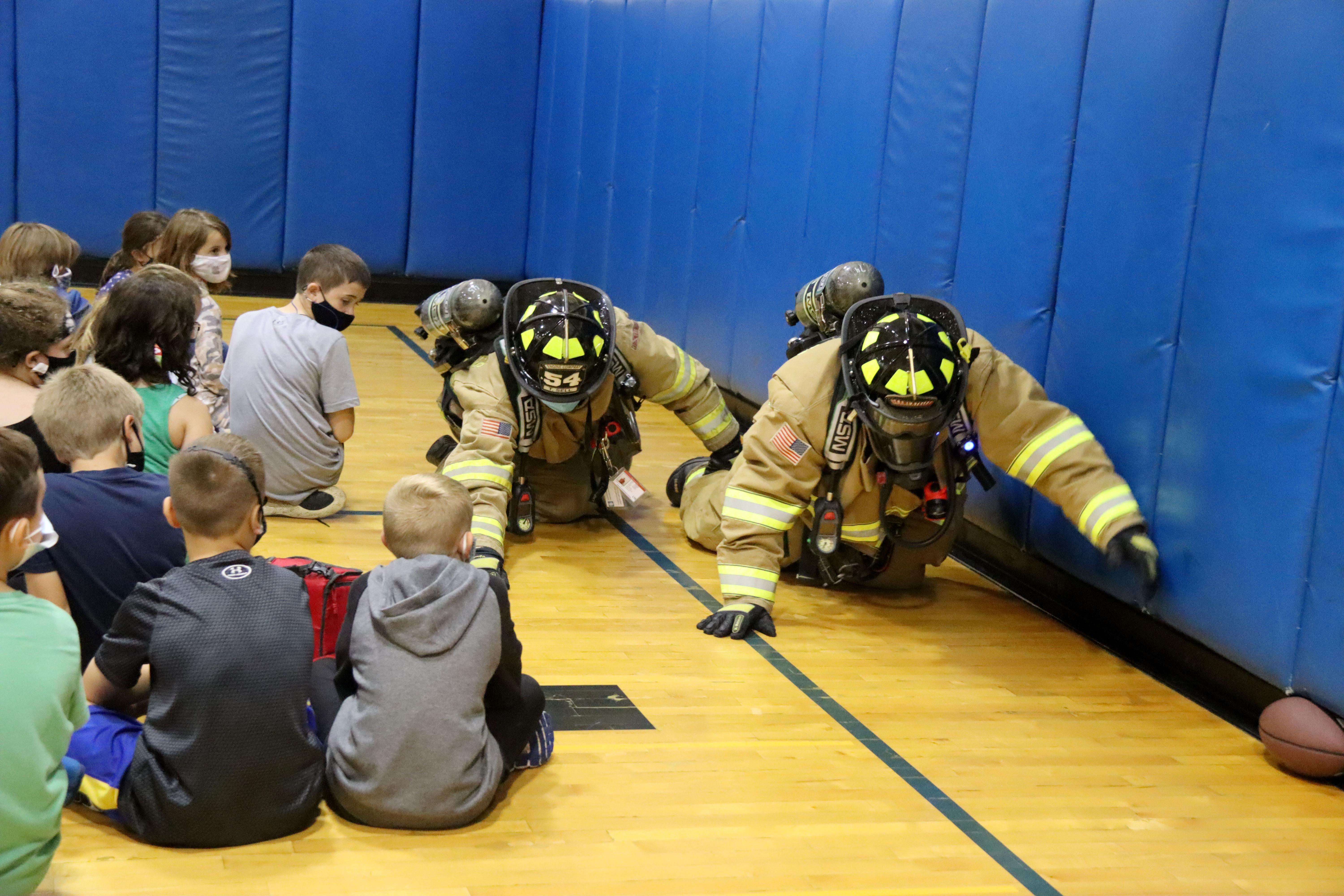 Students look on as firefighters feel the wall as they crawl to the doorway